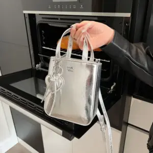 Kleine silberne Henkeltasche mit Herzanhänger, in der Hand vor einem Backofen gehalten