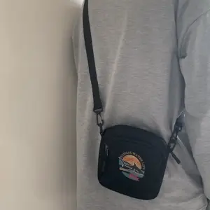 Kleine schwarze Stoff-Umhängetasche mit aufgedrucktem Motiv, getragen vor grauem Pullover
